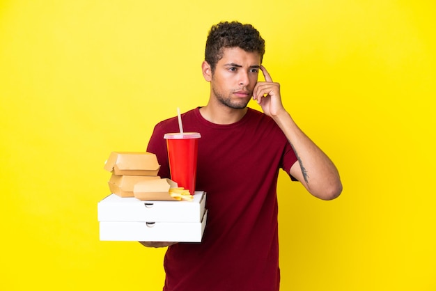 Jonge Braziliaanse man met pizza's en hamburgers geïsoleerde achtergrond met twijfels en denken