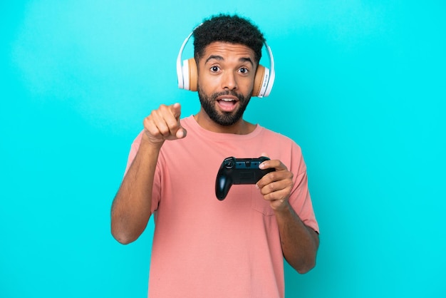 Jonge Braziliaanse man spelen met een video game controller geïsoleerd op blauwe achtergrond verrast en wijzend naar voren
