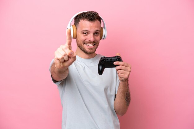 Jonge Braziliaanse man spelen met video game controller geïsoleerd op roze achtergrond tonen en optillen van een vinger