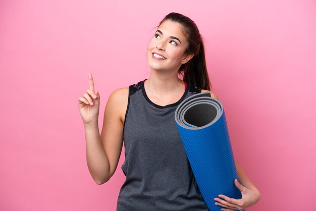 Jonge Braziliaanse sportvrouw die naar yogalessen gaat terwijl ze een mat vasthoudt die op een roze achtergrond is geïsoleerd en een geweldig idee naar boven wijst