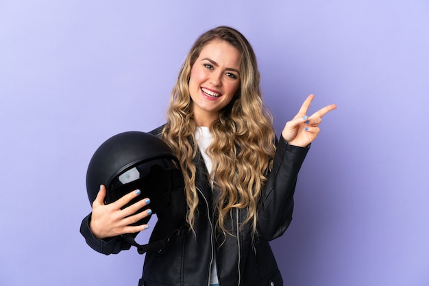 Jonge Braziliaanse vrouw die een motorhelm houdt die op purpere achtergrond wordt geïsoleerd die en overwinningsteken glimlacht toont