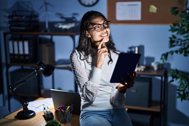 Jonge braziliaanse vrouw die 's nachts touchpad gebruikt en op kantoor werkt met hand op kin na te denken over vraag, peinzende uitdrukking. lachend en attent gezicht. twijfel concept.