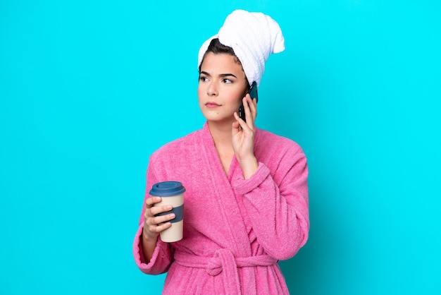 Jonge Braziliaanse vrouw met een badjas geïsoleerd op een blauwe achtergrond met koffie om mee te nemen en een mobiel