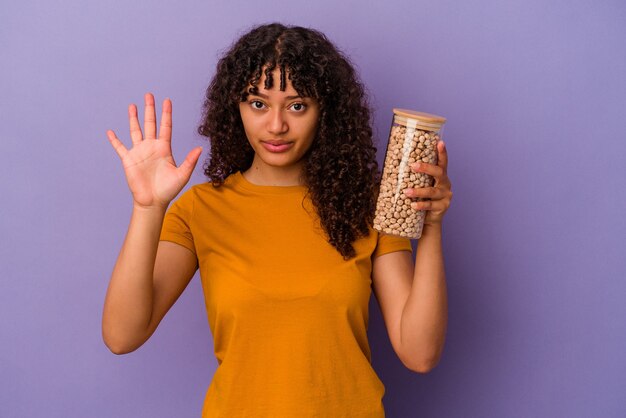 Jonge braziliaanse vrouw met een fles kikkererwten geïsoleerd op paarse achtergrond glimlachend vrolijk nummer vijf met vingers.