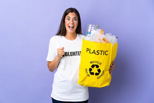 Jonge Braziliaanse vrouw met een zak vol plastic flessen om te recyclen op paars en viert een overwinning in de winnaarspositie