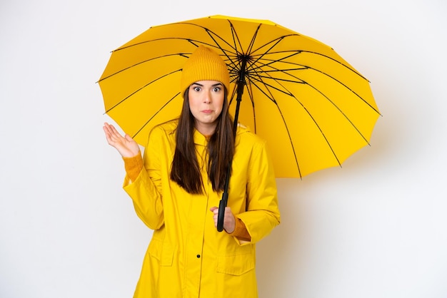 Jonge Braziliaanse vrouw met regenbestendige jas en paraplu geïsoleerd op een witte achtergrond die twijfels heeft terwijl ze de handen opsteekt