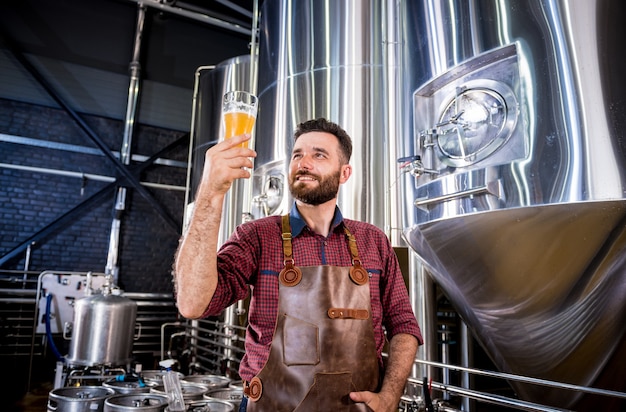 Foto jonge brouwer die een leren schort draagt, test bier in een moderne brouwerij