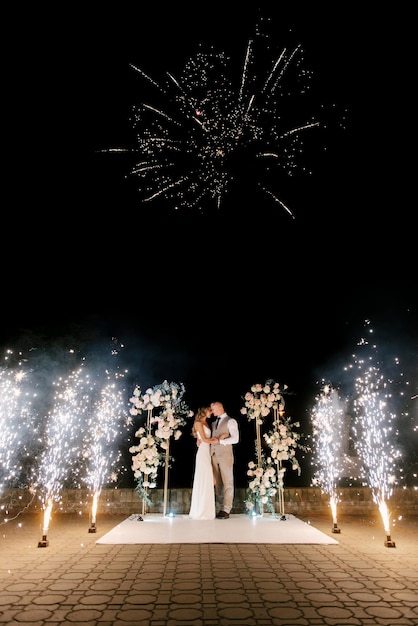 Jonge bruid en bruidegom staan 's nachts in de buurt van de huwelijksboog met lichten, rook en vuurwerksilhouetten van de pasgetrouwden
