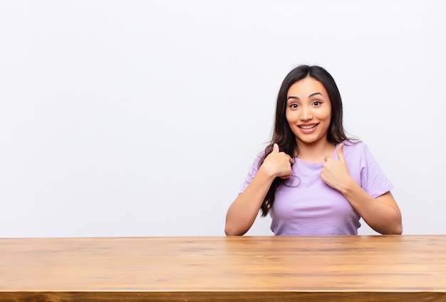 Jonge brunette achter een bureau verrast en trots wijzend naar zichzelf met een opgewonden verbaasde blik