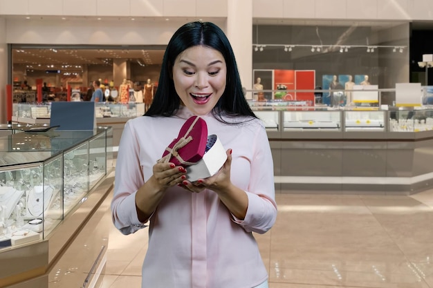Jonge brunette aziatische vrouw opent een hartvormige geschenkdoos gezichtsuitdrukking van het interieur van een verrassingswinkelcentrum