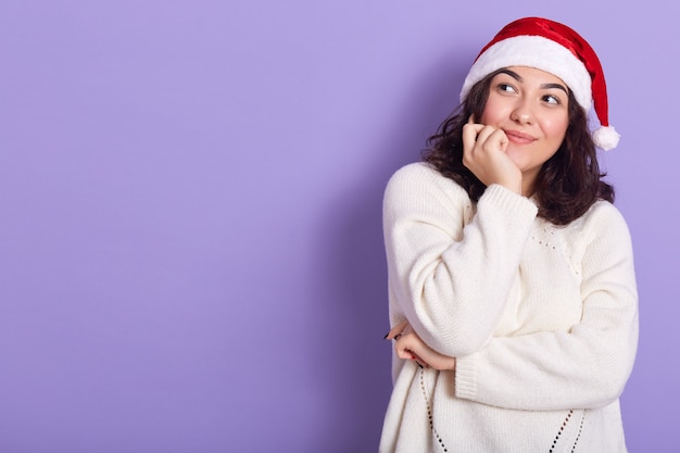 Jonge brunette die opzij kijkt, wat betreft haar kin met hand, die witte sweater en de rode hoed van de Kerstman draagt