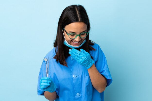 Jonge brunette gemengd ras tandarts vrouw met tools over geïsoleerde muur gelukkig en lachend die mond met handen