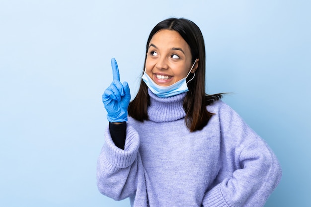 Jonge brunette gemengd ras vrouw beschermen tegen het coronavirus met een masker en handschoenen over geïsoleerde blauwe muur die een geweldig idee