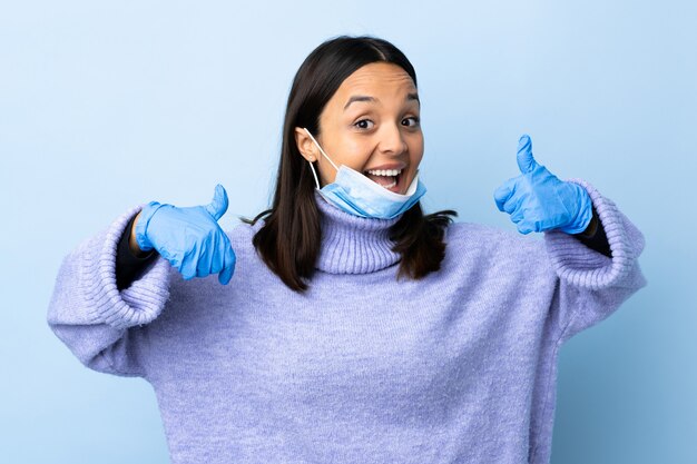 Jonge brunette gemengd ras vrouw beschermen tegen het coronavirus met een masker en handschoenen over geïsoleerde blauwe muur met een thumbs up gebaar