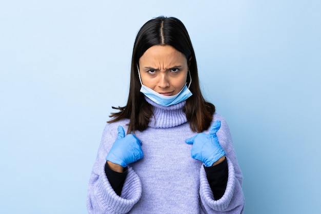 Jonge brunette gemengd ras vrouw beschermen tegen het coronavirus met een masker en handschoenen over geïsoleerde blauwe muur naar zichzelf