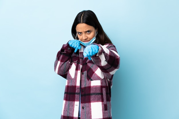 Jonge brunette gemengd ras vrouw bescherming tegen het coronavirus met een masker en handschoenen over geïsoleerde blauwe muur met vechten gebaar