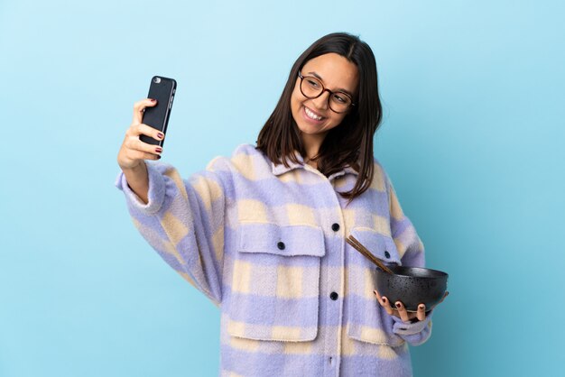 Jonge brunette gemengd ras vrouw met een kom vol noedels over geïsoleerde blauwe muur maken van een selfie