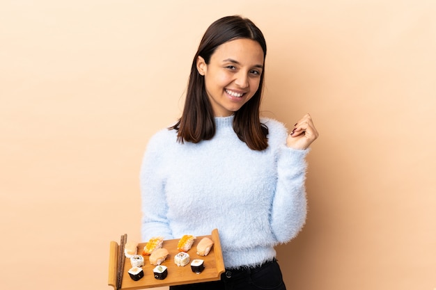 Jonge brunette gemengd ras vrouw met sushi over muur lachen