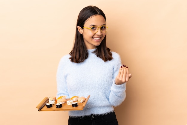 Jonge brunette gemengd ras vrouw met sushi over muur uitnodigen om met de hand te komen. Blij dat je bent gekomen