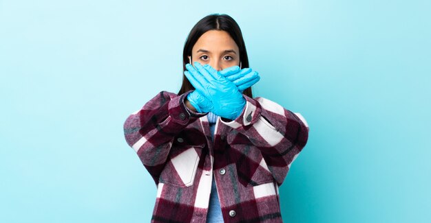Jonge brunette gemengde rasvrouw die met een masker en handschoenen over blauwe muur beschermen die eindegebaar met haar hand maken