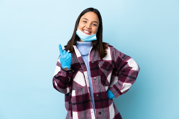 Jonge brunette gemengde rasvrouw die met een masker en handschoenen over blauwe muur beschermen die komend gebaar doen