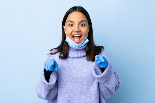 Jonge brunette gemengde rasvrouw die met een masker en handschoenen over blauwe muur beschermt verrast en voorzijde richt