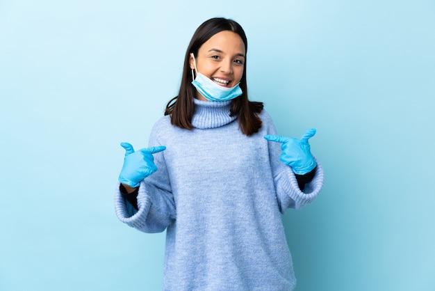 Jonge brunette gemengde rasvrouw die met een masker en handschoenen over blauwe trots en zelfverzekerde muur beschermen