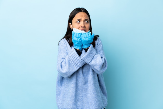 Jonge brunette gemengde rasvrouw die met een masker en handschoenen over blauwe zenuwachtige en doen schrikken muur beschermen die handen op mond zetten