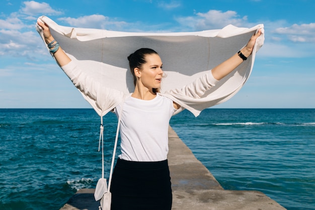 Jonge brunette in een vest en een rok die zich op een pijler op de achtergrond van het overzees bevinden en geniet van een de zomerwind op een zonnige dag