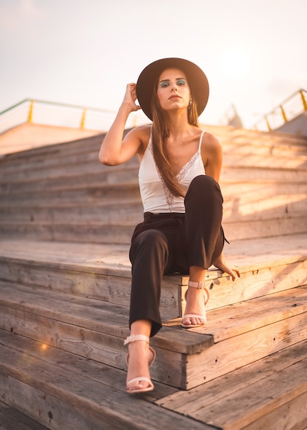 Foto jonge brunette in een zwarte hoed en een wit t-shirt genieten van zomer zittend op een houten trap