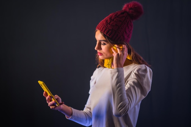 Jonge brunette kaukasisch met gele koptelefoon, wit t-shirt en wollen hoed op een zwarte achtergrond, kijkend naar de mobiele telefoon
