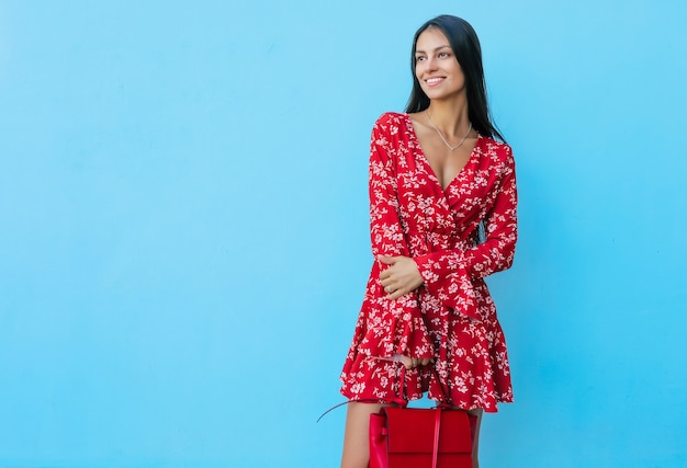 Jonge brunette meisje poseert op een blauwe achtergrond in een rode jurk, houdt een rode handtas vast, glimlacht en kijkt naar links