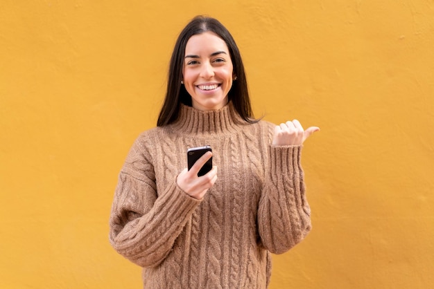 Jonge brunette vrouw buitenshuis met behulp van mobiele telefoon en wijzend naar de zijkant