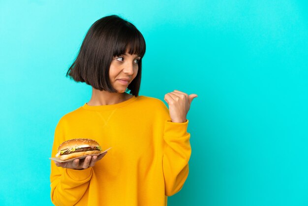 Jonge brunette vrouw die een hamburger over een geïsoleerde achtergrond vasthoudt en naar de zijkant wijst om een product te presenteren