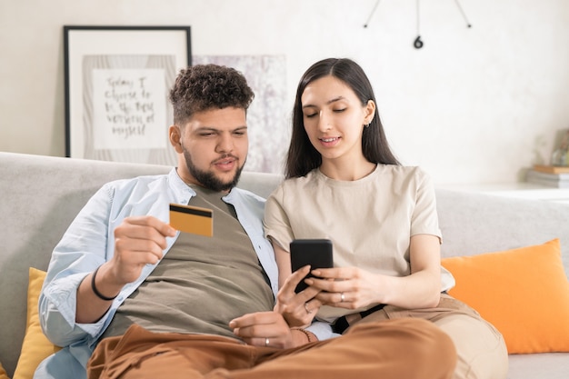 Jonge brunette vrouw die lekker eten in smartphone toont aan haar man met creditcard terwijl ze lunch gaat bestellen in online café