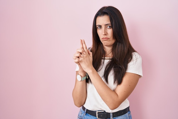 Jonge brunette vrouw die over roze achtergrond staat en symbolisch pistool met handgebaar vasthoudt, dodende schietwapens speelt, boos gezicht