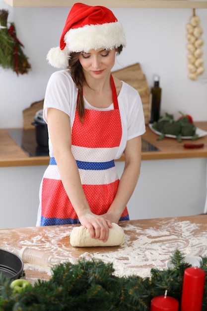 Jonge brunette vrouw die pizza of handgemaakte pasta kookt terwijl ze een kerstmuts in de keuken draagt. Huisvrouw bereidt deeg op houten tafel. Gelukkig nieuwjaar, trouwen met kerst, diëten, eten en gezondheid