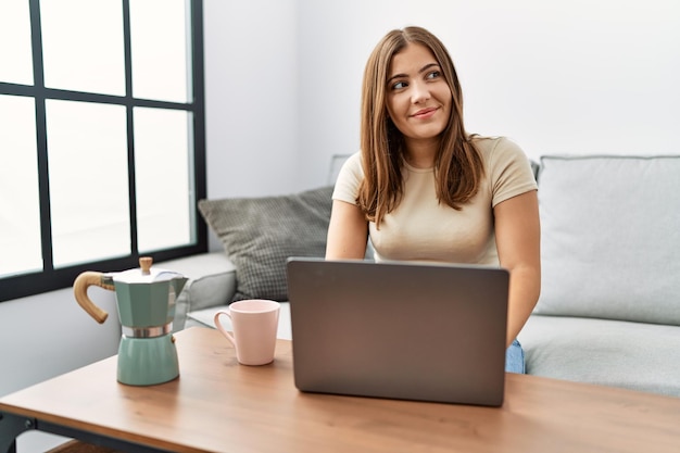 Jonge brunette vrouw die thuis een laptop gebruikt die een kopje koffie drinkt glimlachend naar de zijkant kijkend en weg starend denkend