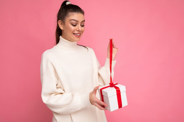 jonge brunette vrouw geïsoleerd op roze achtergrond muur dragen witte trui bedrijf geschenkdoos en unboxing aanwezig kijken doos.