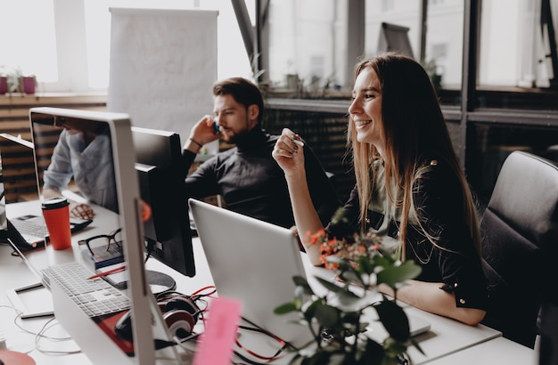 Jonge brunette vrouw gekleed in kleding in kantoorstijl werkt op de computer aan het bureau met de collega's in het moderne kantoor