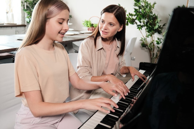 Jonge brunette vrouw haar slimme tienerdochter raadplegen terwijl zowel zittend door piano als samen spelen in de thuisomgeving