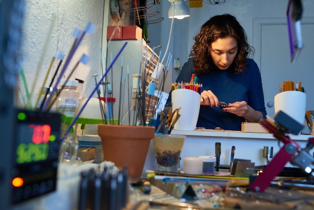 Jonge brunette vrouw in blauwe trui omgeven door leveringen zitten door tafel in werkplaats en werkstukken maken voor verder werk