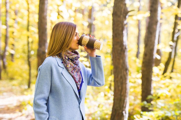 Jonge brunette vrouw in een blauwe jas staan in het park met een kopje koffie