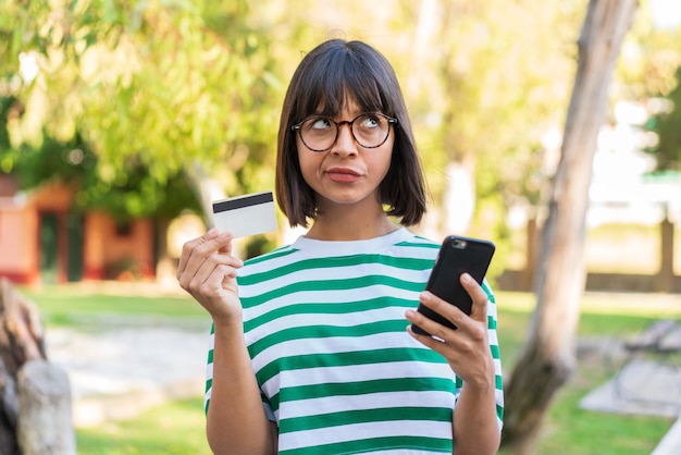 Jonge brunette vrouw in het park kopen met de mobiel met een creditcard terwijl ze denken