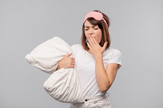 Jonge brunette vrouw in nachtkleding en masker voor haar mond terwijl geeuwen geïsoleerd