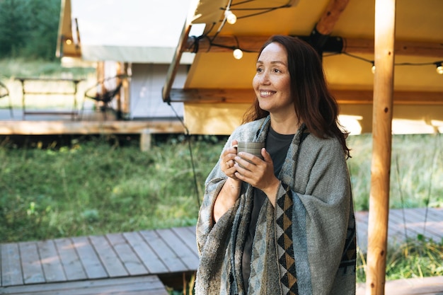 Jonge brunette vrouw in poncho thee drinken en ontspannen in glamping in de natuur