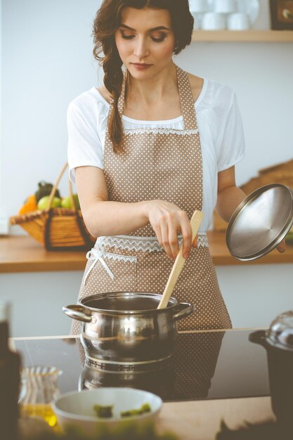 Jonge brunette vrouw kokende soep in de keuken. Huisvrouw die houten lepel in haar hand houdt. Voedsel en gezondheidsconcept.