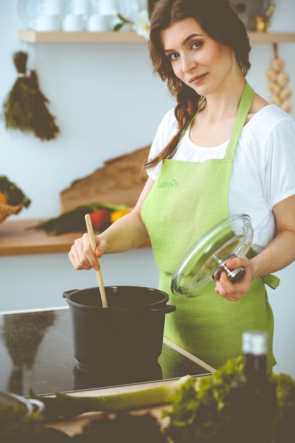 Jonge brunette vrouw kokende soep in de keuken. Huisvrouw die houten lepel in haar hand houdt. Voedsel en gezondheidsconcept.