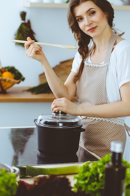 Foto jonge brunette vrouw kokende soep in de keuken. huisvrouw die houten lepel in haar hand houdt. voedsel en gezondheidsconcept.