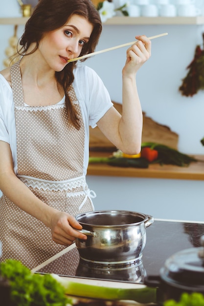 Jonge brunette vrouw kokende soep in de keuken. Huisvrouw die houten lepel in haar hand houdt. Voedsel en gezondheidsconcept.